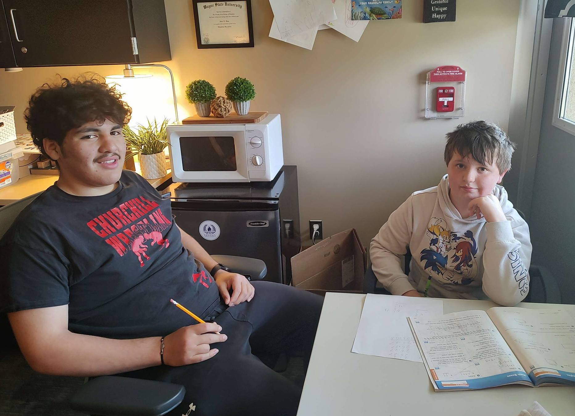 Two boys occupy a small study space, one leaning back with a pencil in hand and the other resting his chin on his hand while working at a table with open worksheets and a magazine.
