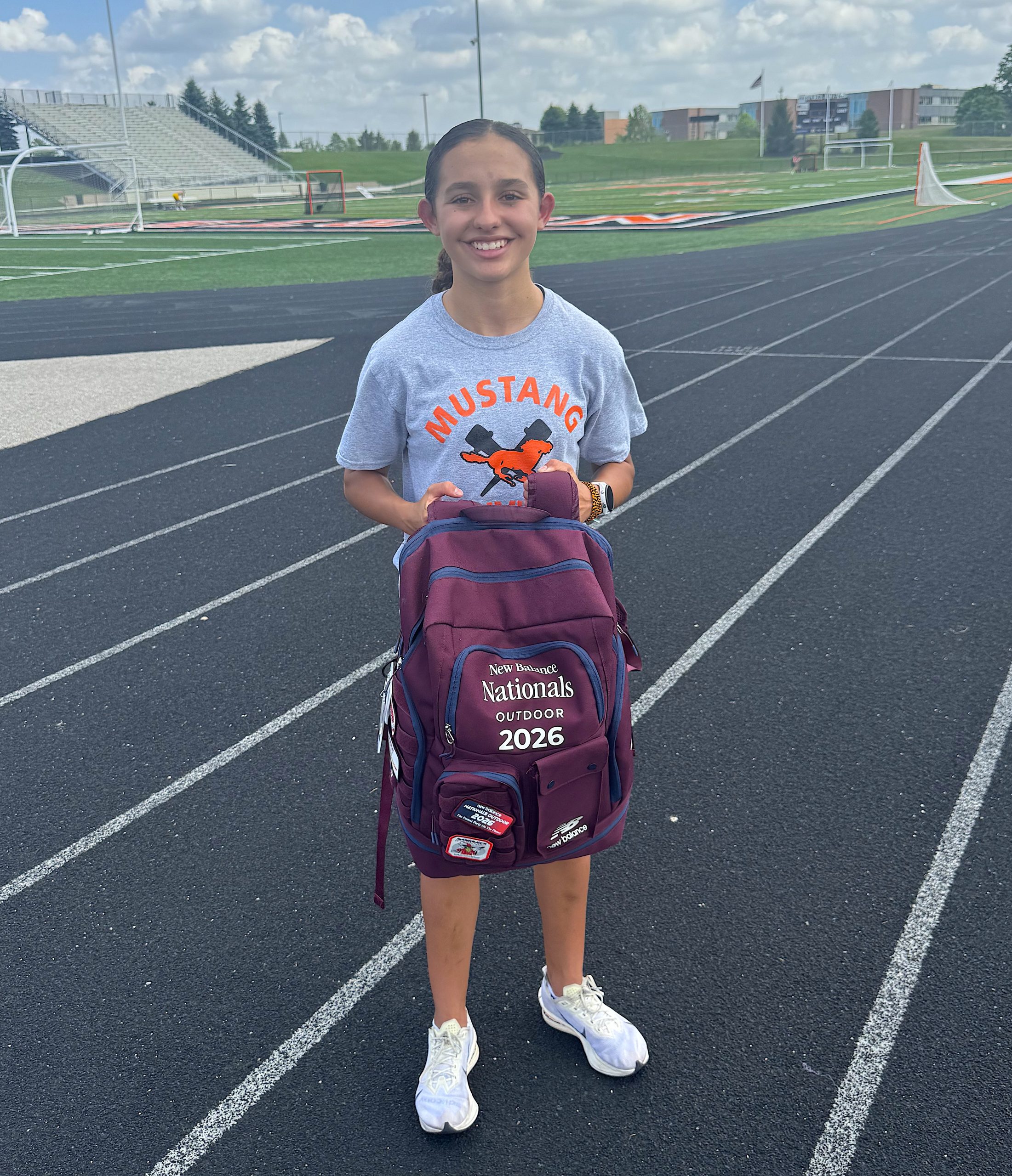 Alex1 - Social House News Smiling young girl on a track field, wearing a gray Mustang T-shirt and a maroon backpack, outdoors on a running track.
