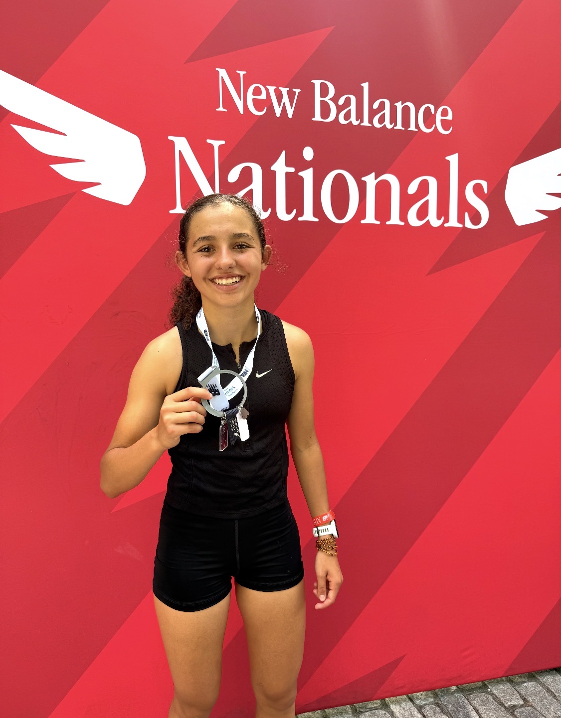 Screenshot - Social House News Smiling young female athlete in a black athletic outfit holding medals in front of a red New Balance Nationals backdrop.