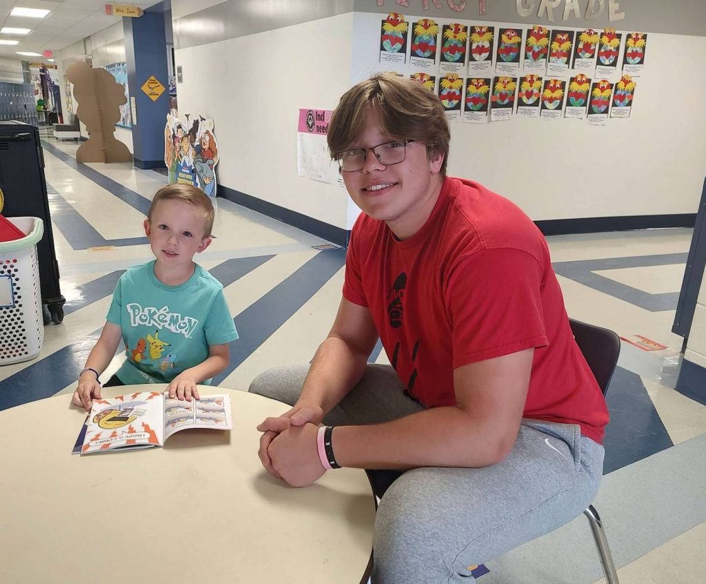 Teenager with glasses in a red shirt sits beside a young boy at a round table in a school hallway, reading a comic book together.
