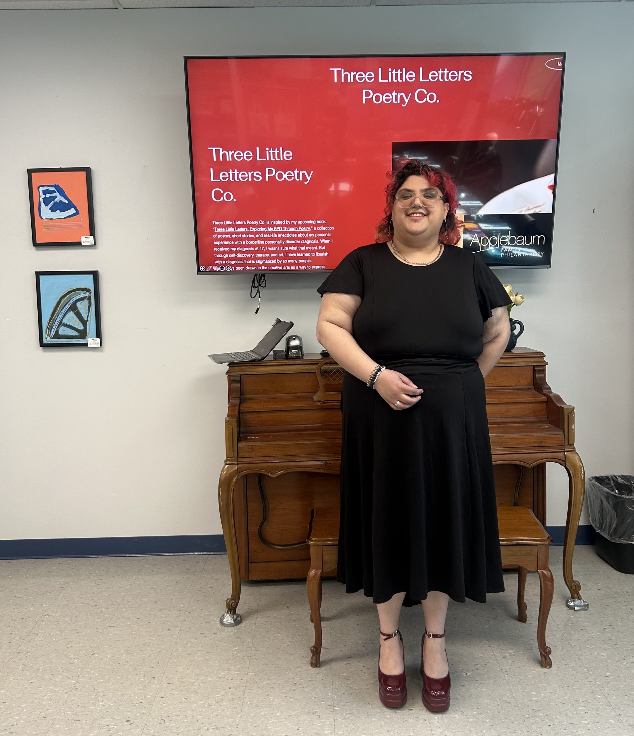 Creative Expressions poet Jo Banooni - Social House News Smiling woman in a black dress stands beside a wooden piano in a gallery, with a large red screen showing 'Three Little Letters Poetry Co.' behind her.