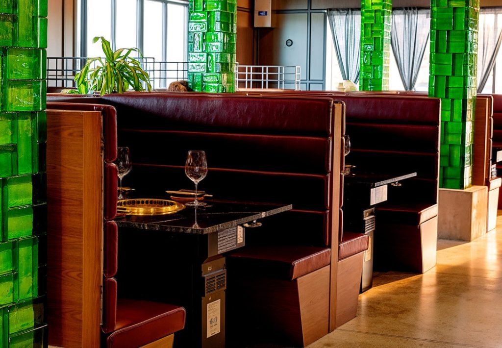 Restaurant booths with burgundy leather seating, green tiled columns, and wine glasses on the table by large windows.