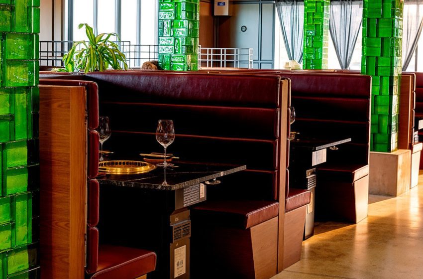 Restaurant booths with burgundy leather seating, green tiled columns, and wine glasses on the table by large windows.