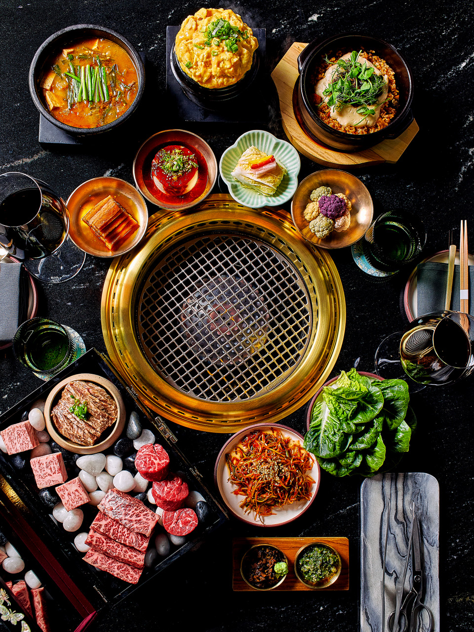 Top-down view of a round grill surrounded by assorted bowls and plates of beef, vegetables, and side dishes for a yakiniku meal, on a dark countertop.