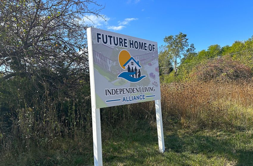 FuturehomeILA - Social House News Outdoor sign reading 'Future Home of Independent Living Alliance' with a logo of a house on a blue wave and sun, mounted on two posts in a grassy area.
