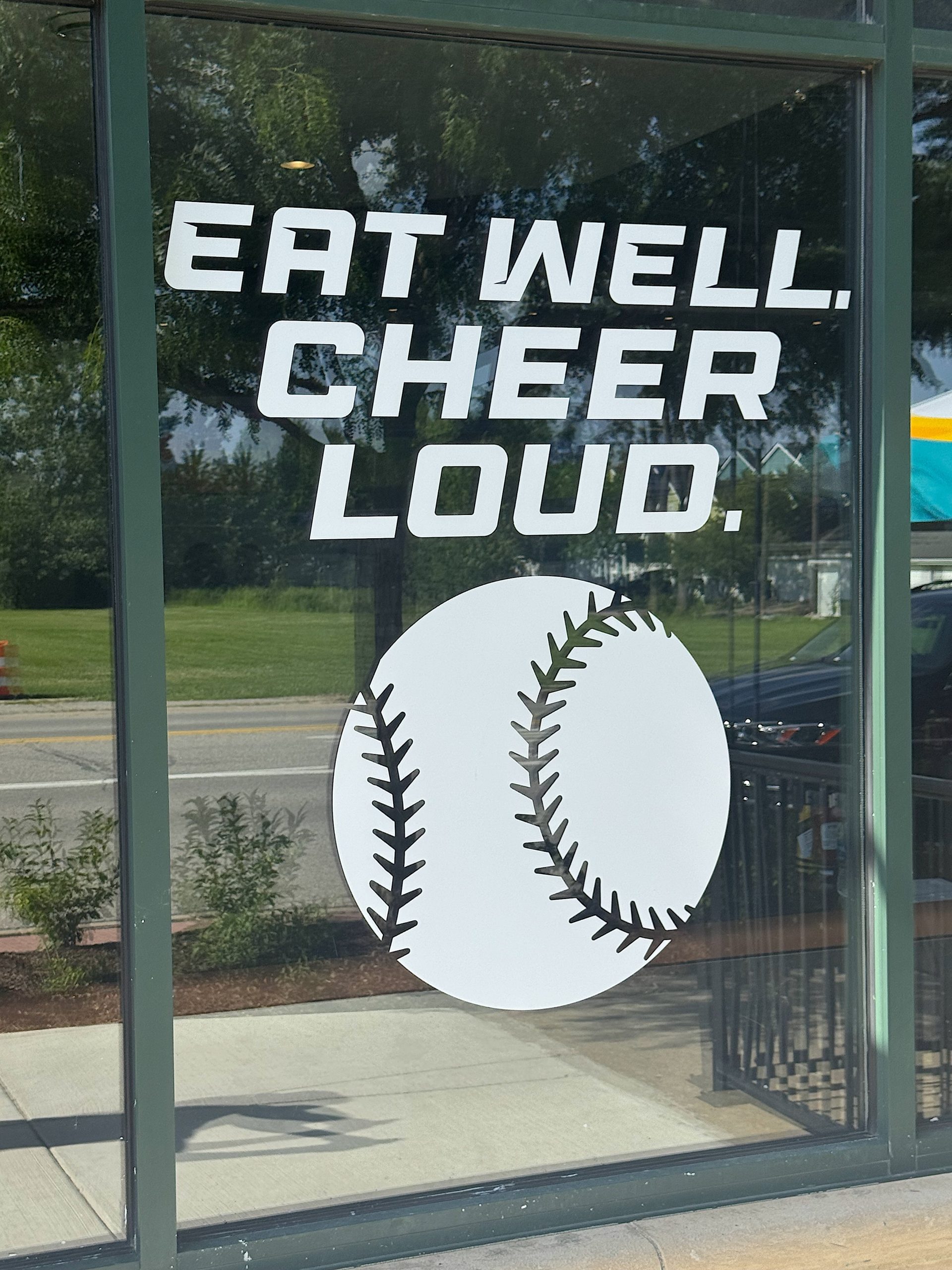 Sign on a glass storefront showing the slogan 'EAT WELL. CHEER LOUD.' alongside a large white baseball with black stitches beneath it.