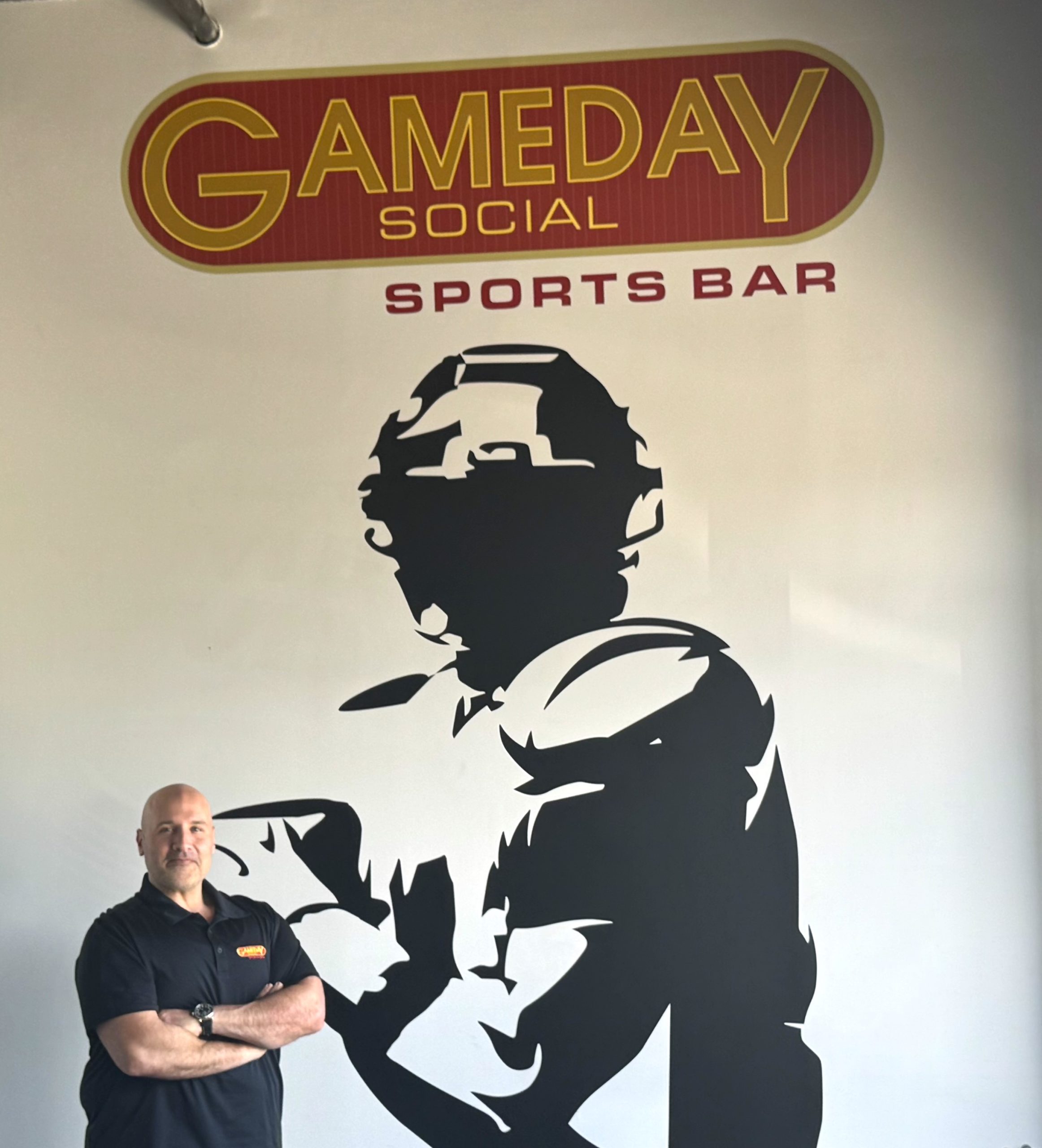 Man standing with arms crossed in front of a wall mural featuring a large black silhouette of a person and a 'GAME DAY Social Sports Bar' sign above.