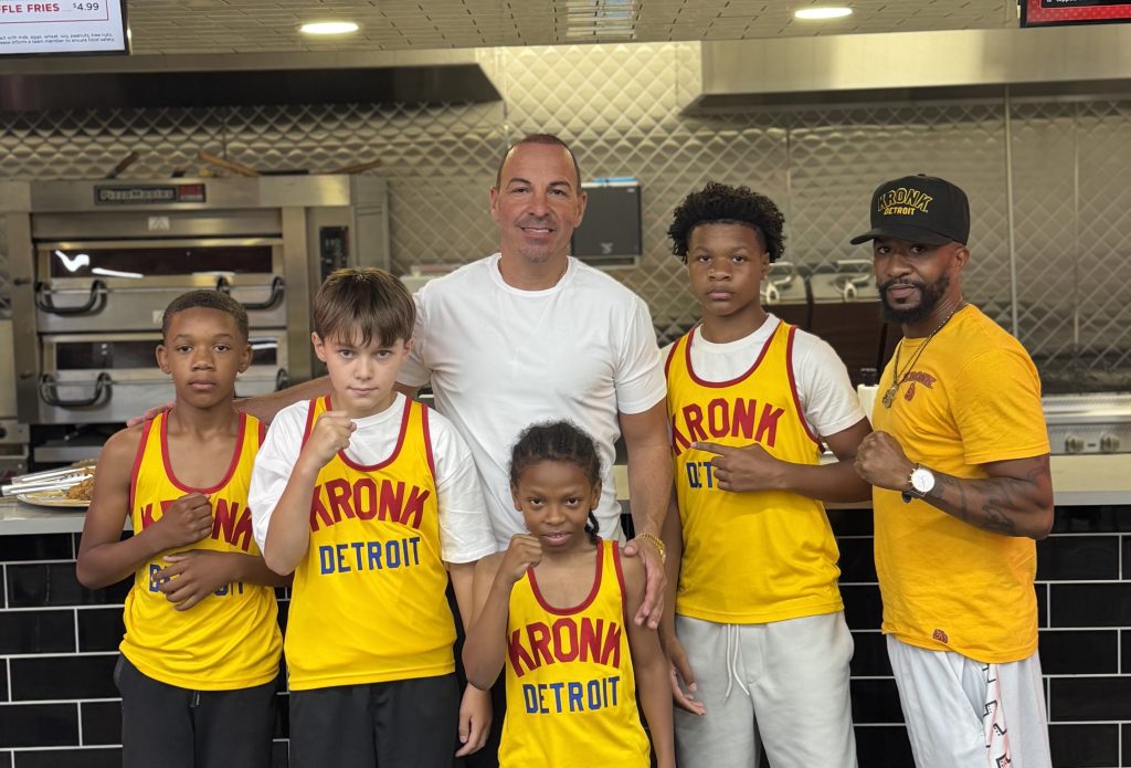 Group of Kronk Detroit basketball players and coaches posing together in a stainless steel kitchen, wearing yellow uniforms with red lettering.