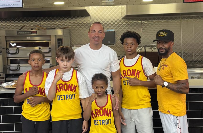 Group of Kronk Detroit basketball players and coaches posing together in a stainless steel kitchen, wearing yellow uniforms with red lettering.