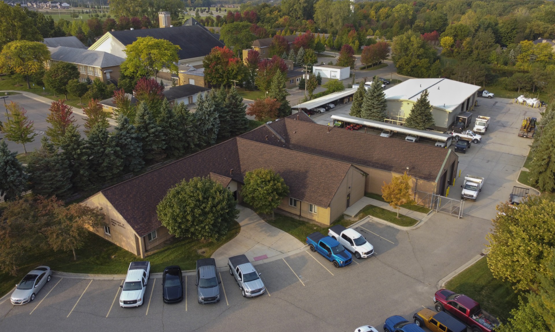 Headquarters - Social House News Aerial view of a campus-like area with a brown-roofed building, surrounding trees, and a parking lot full of cars.