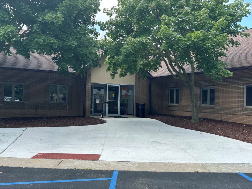 Entrance to a brick building with glass doors shaded by large trees, and a curved concrete walkway in the foreground.