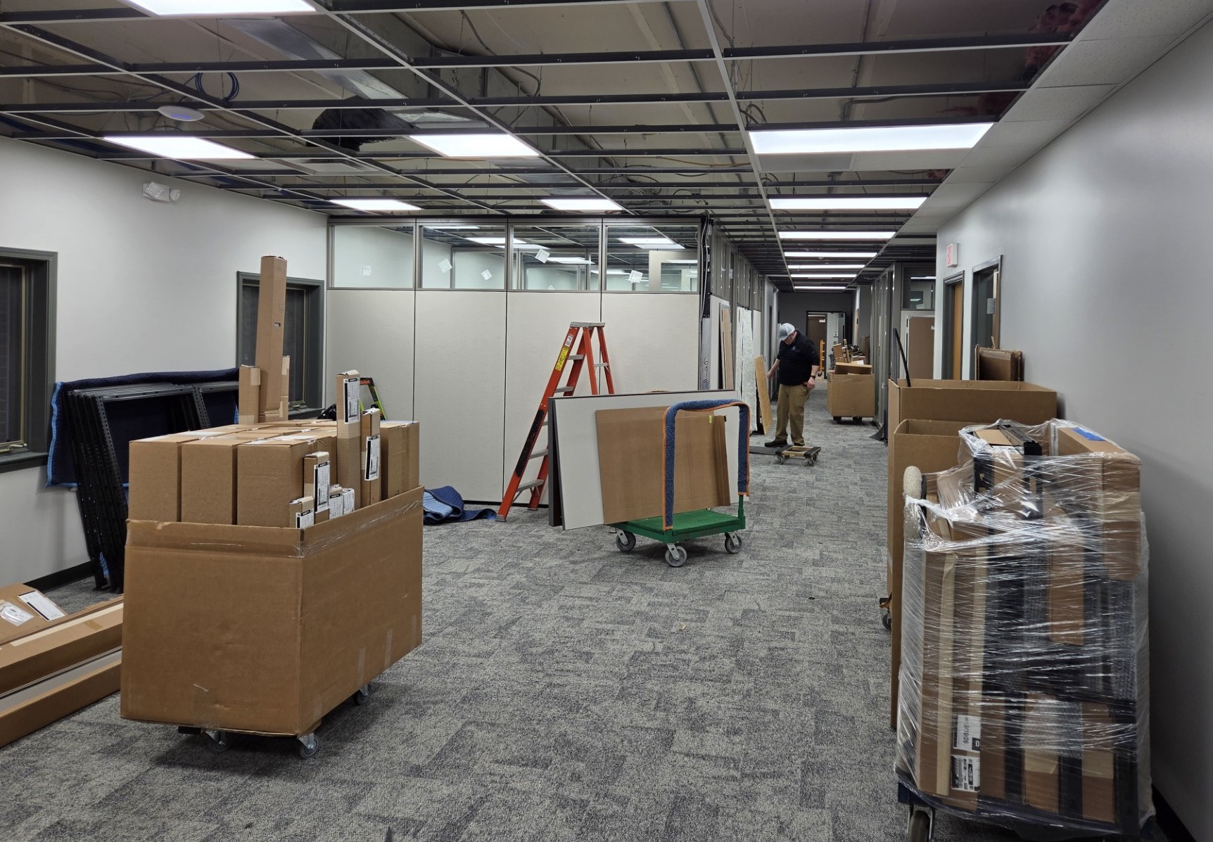 Office hallway under renovation with exposed ceiling grid, ladders, and stacked boxes on carts; a worker stands in the distance.