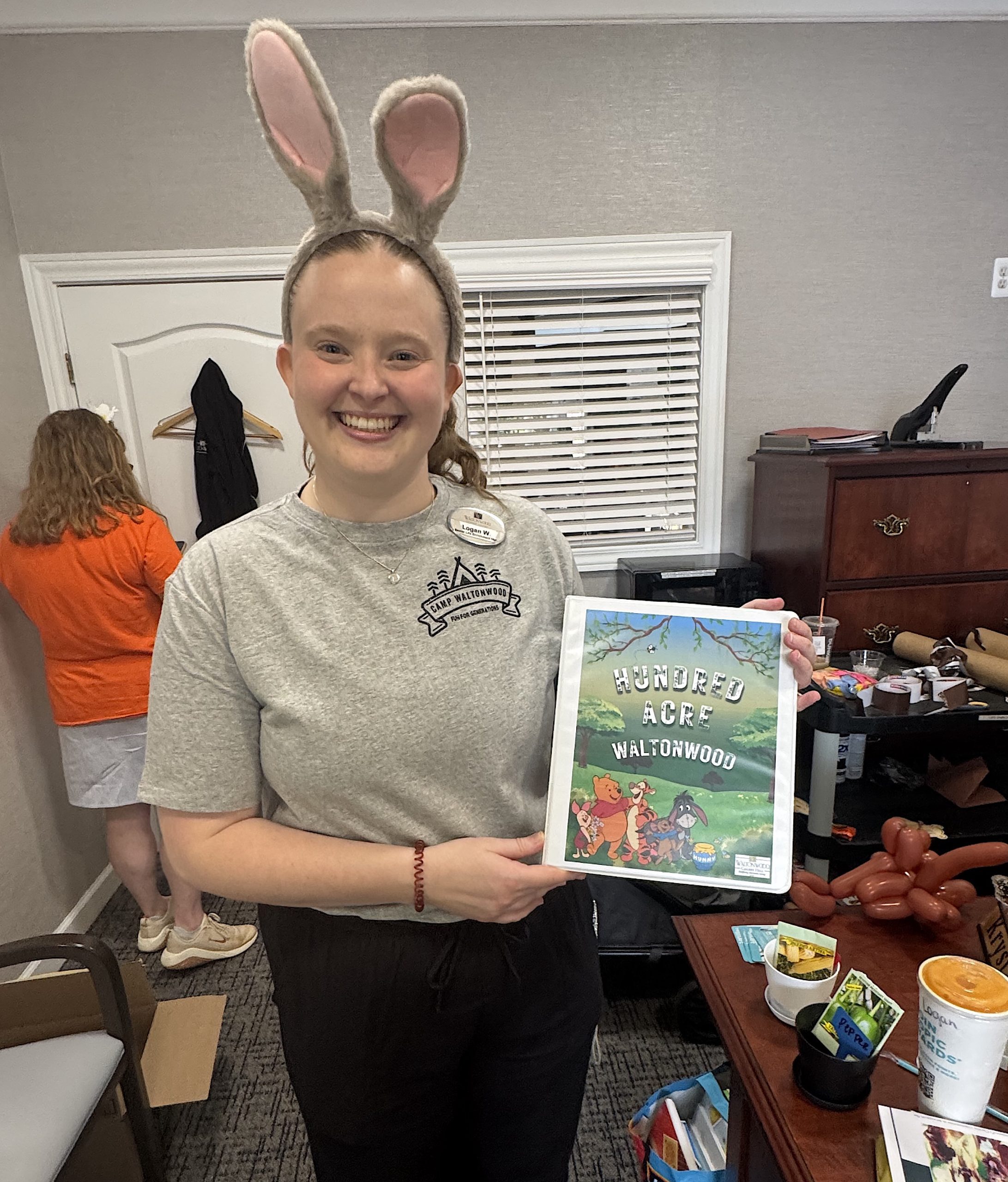 Smiling person wearing fuzzy bunny ears headband, presenting a colorful children's book titled 'Hundred Acre Wood.'