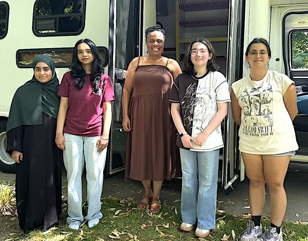 Five diverse women stand together in front of a green bus, smiling for the photo.