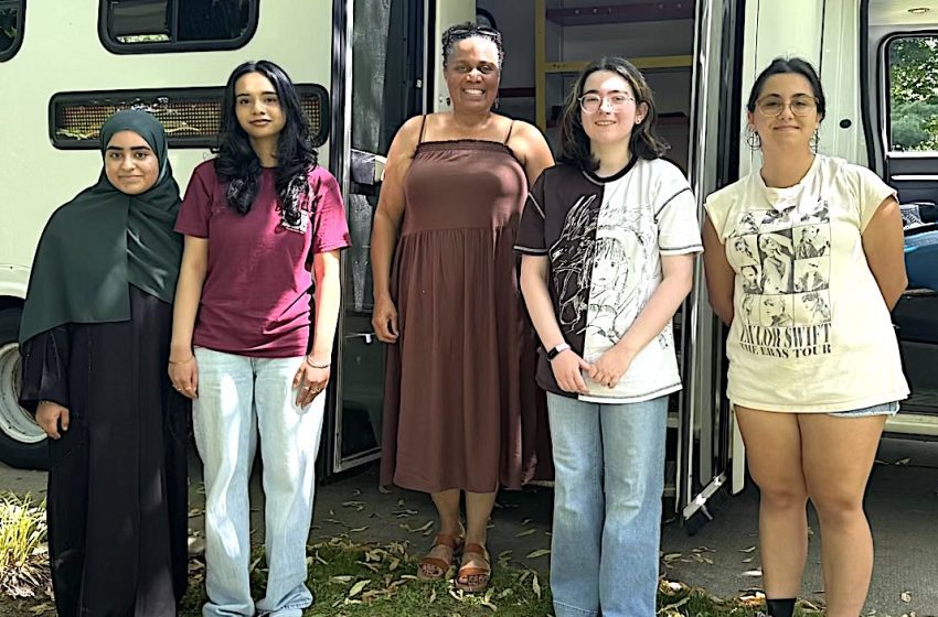 Five diverse women stand together in front of a green bus, smiling for the photo.