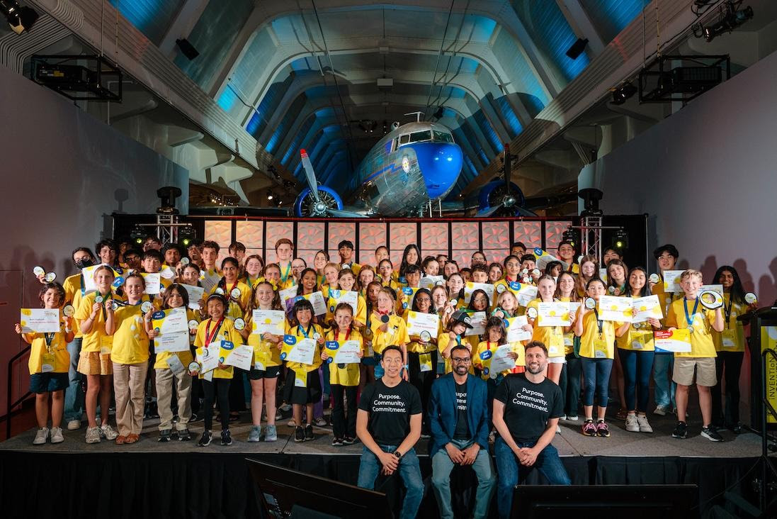 Inventor2 - Social House News Group of children in yellow shirts on a stage, holding certificates and medals at an aviation museum awards ceremony with a large airplane in the background.
