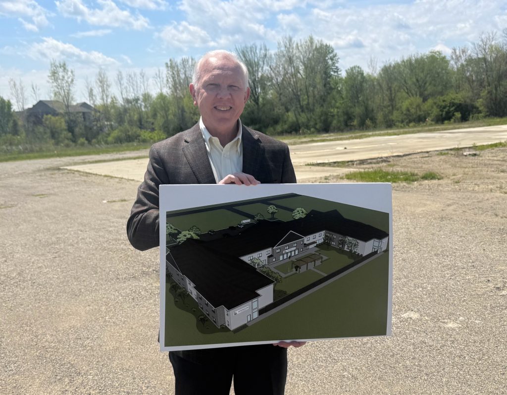 Smiling man in a brown blazer holds a large architectural rendering outdoors on a vacant lot.