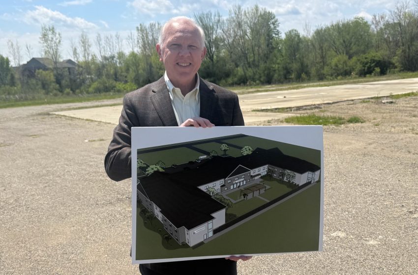 Smiling man in a brown blazer holds a large architectural rendering outdoors on a vacant lot.