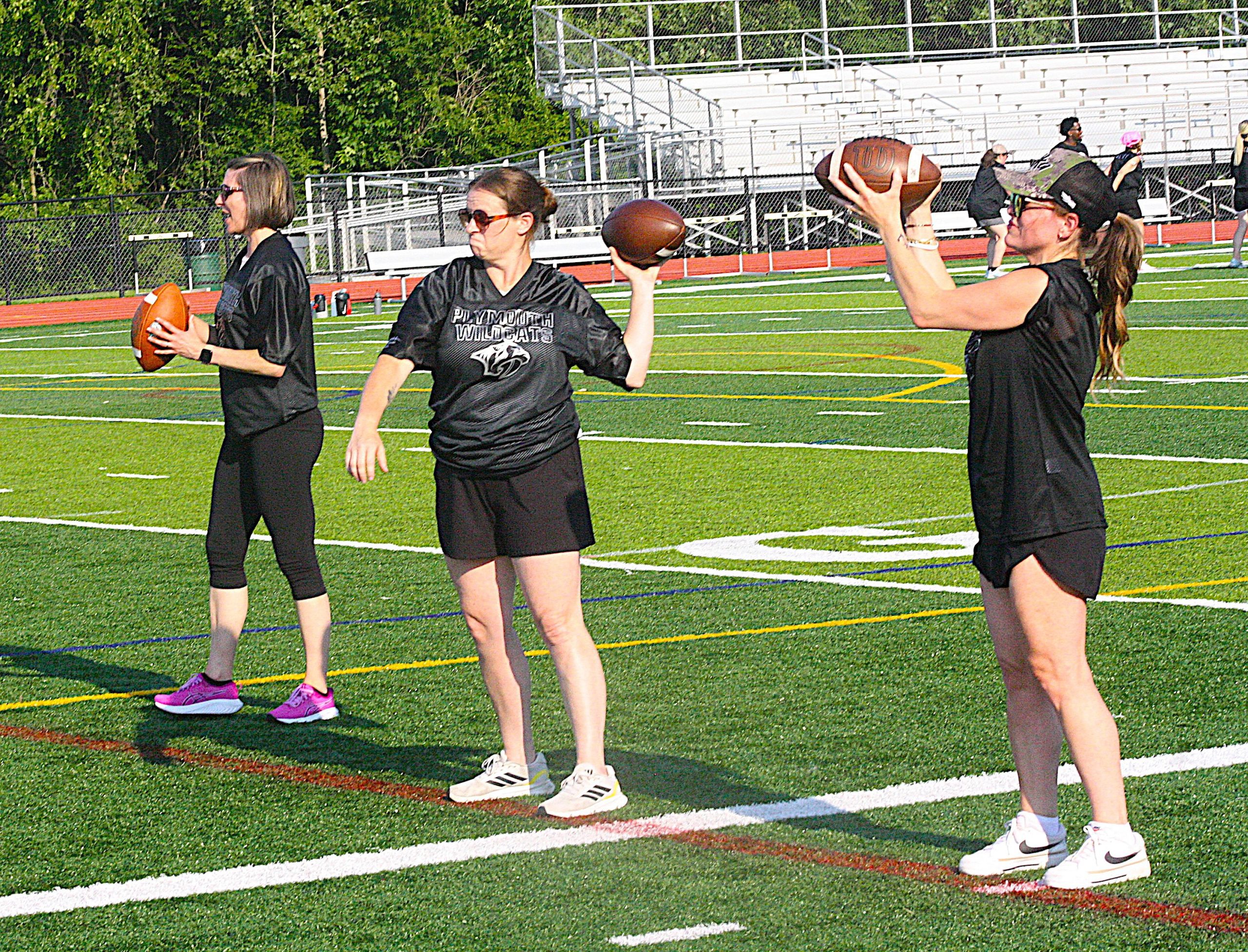 Women on a football field preparing to throw footballs during a practice session on artificial turf.