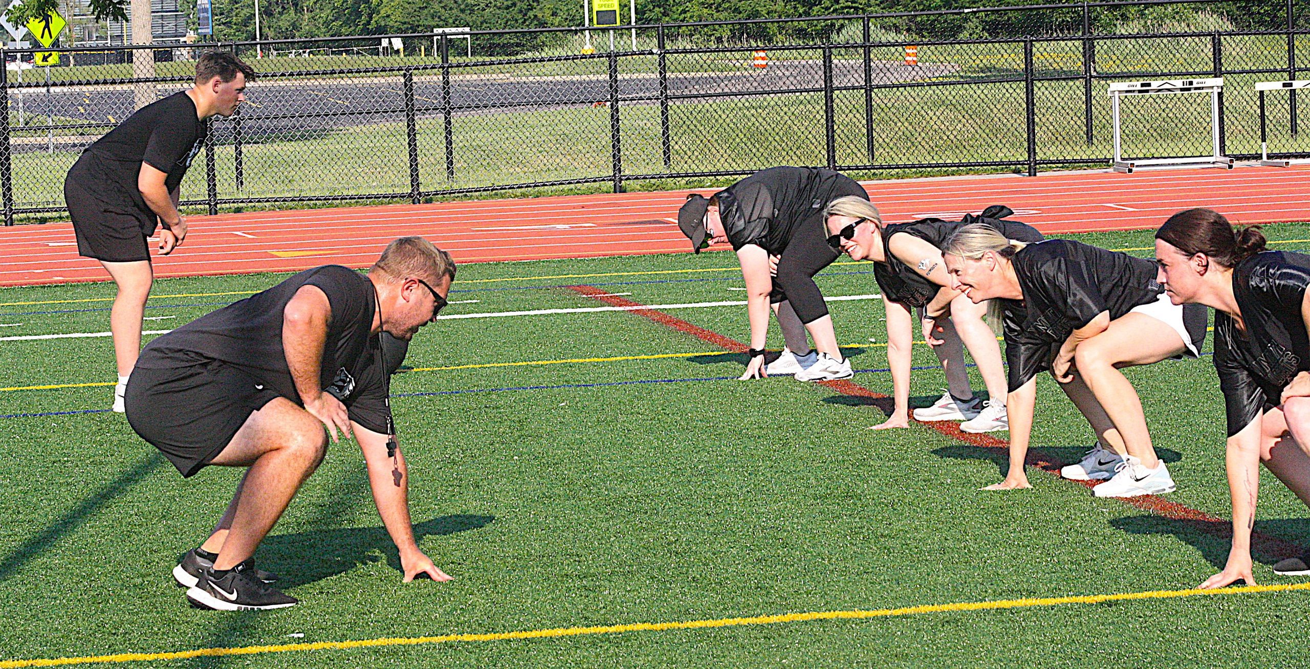 Momstackle8 - Social House News Group of runners in starting crouch on a synthetic track, ready to sprint under a chain-link fence on a sunny day.