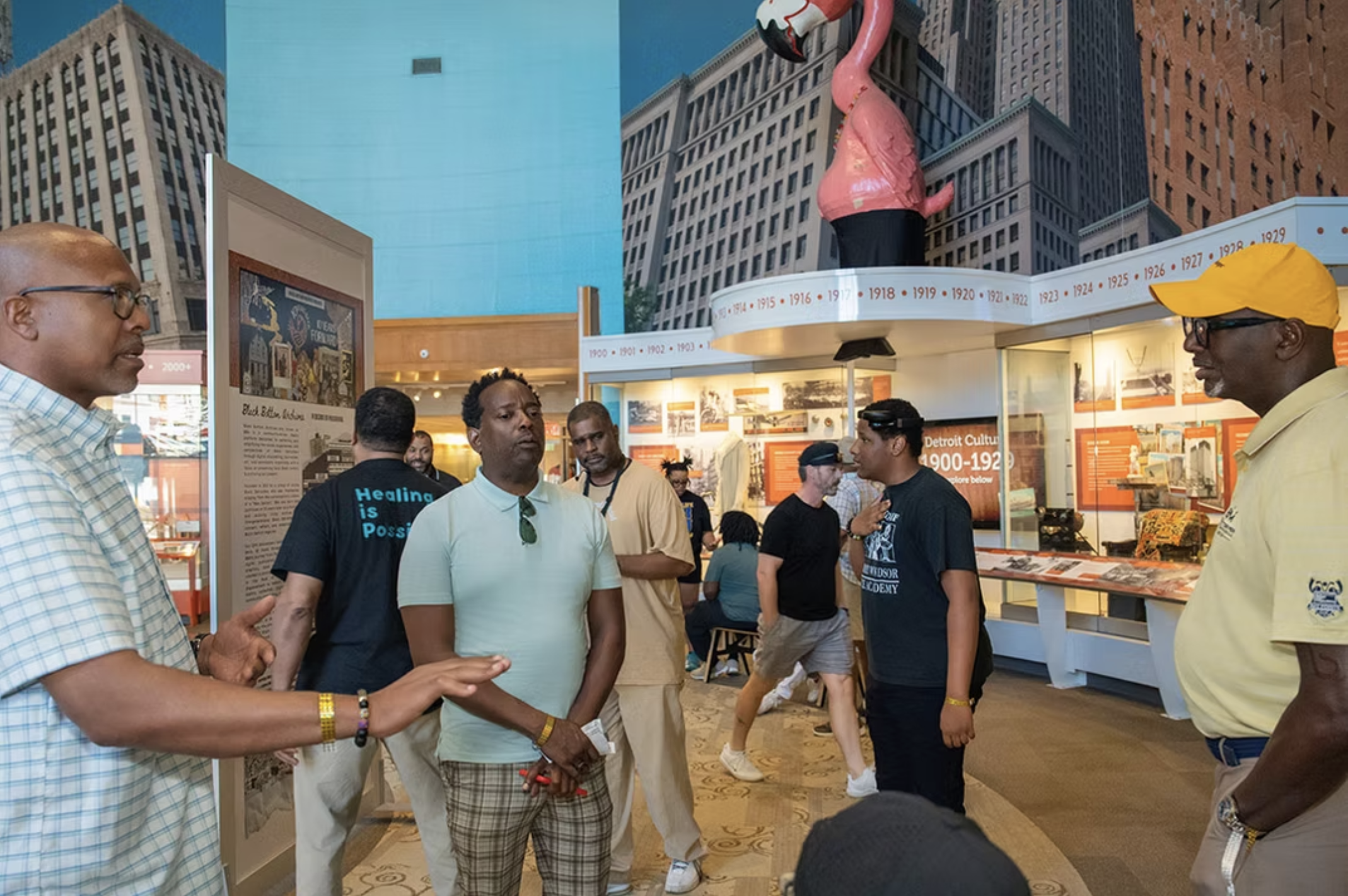 People gather in a museum exhibit with timeline displays and a large pink flamingo statue overhead.