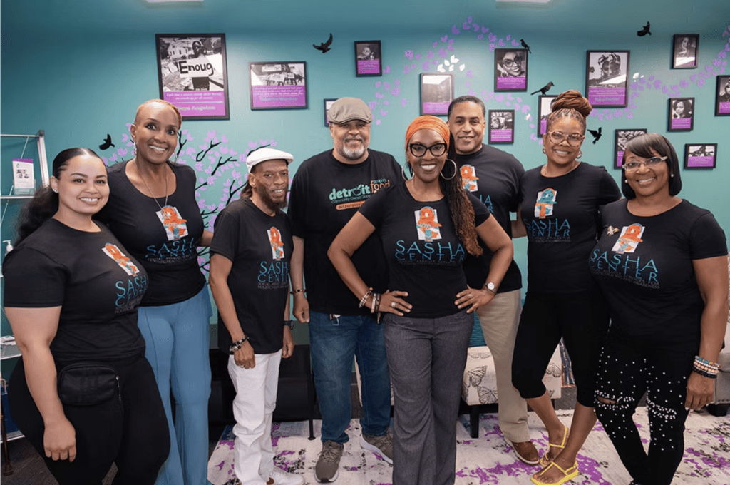 Group of eight adults posing for a photo, wearing black shirts that say SASH Center, in a teal wall room with framed posters and purple decor behind them.