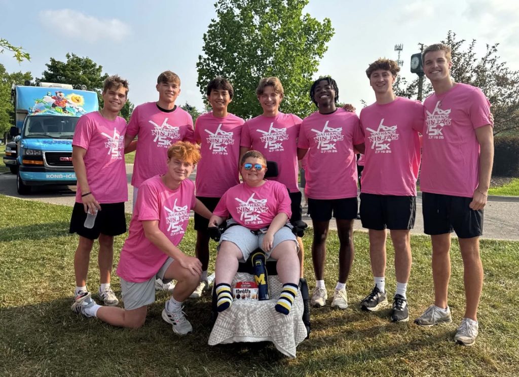 Group of youths in matching pink shirts posing with a child in a wheelchair at an outdoor charity 5K event, smiling for the camera.