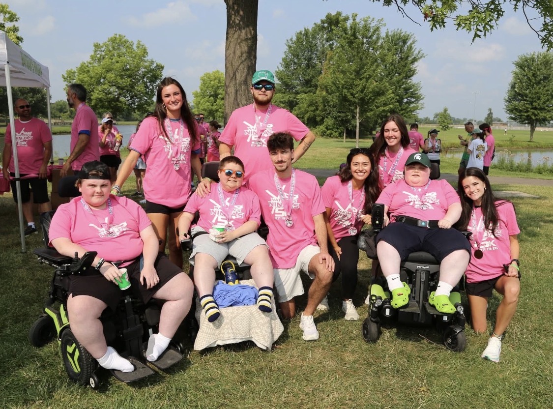 Screenshot - Social House News Group of people wearing matching pink shirts posing for a photo at an outdoor charity event in a park, with several in wheelchairs and medals around their necks.