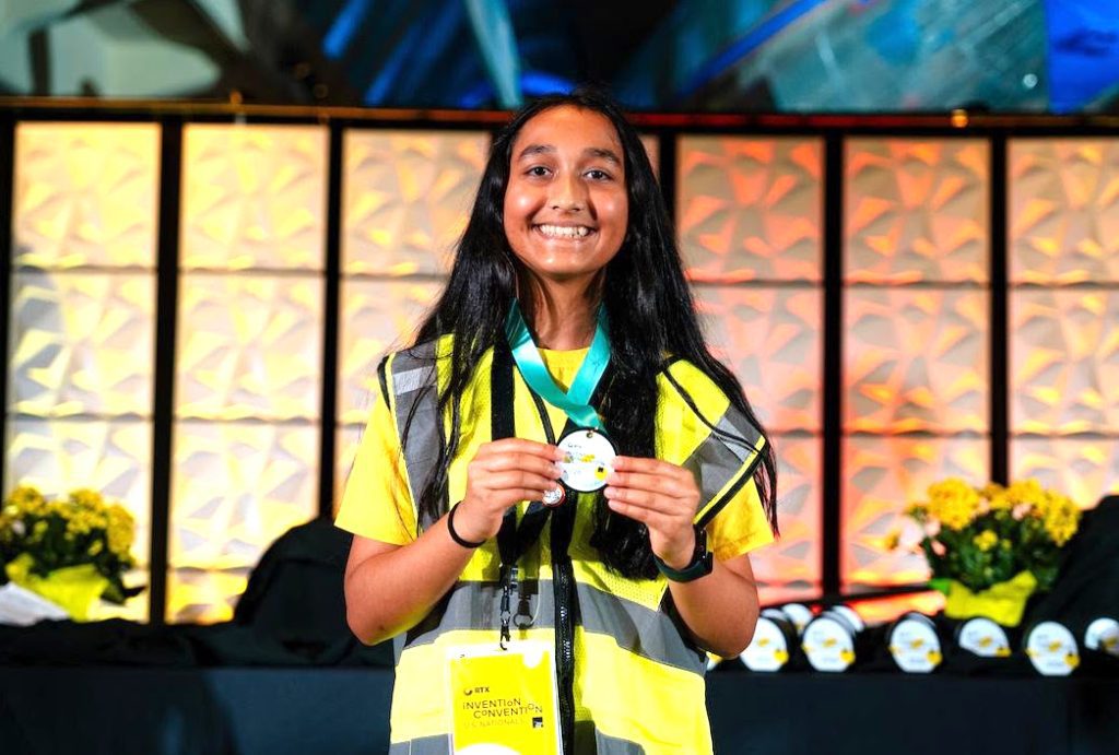 Young woman in a yellow safety vest smiles while holding a medal at an awards ceremony on a stage.