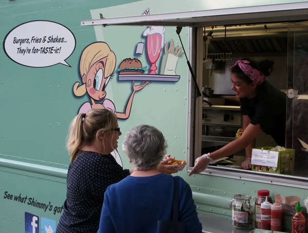 Two customers at a mint-green food truck window receive a burger from a server; the truck features a cartoon waitress and a speech bubble advertising burgers, fries, and shakes.