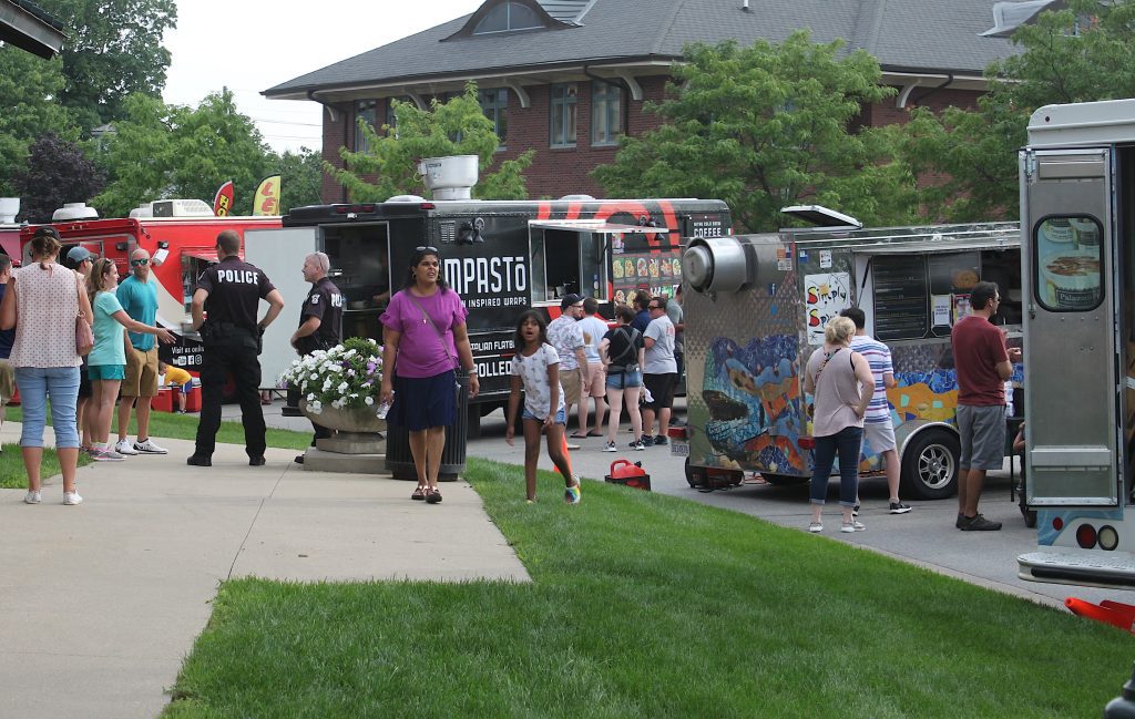 People line up and mingle beside colorful food trucks at a sunny outdoor festival street scene with greenery in the foreground.