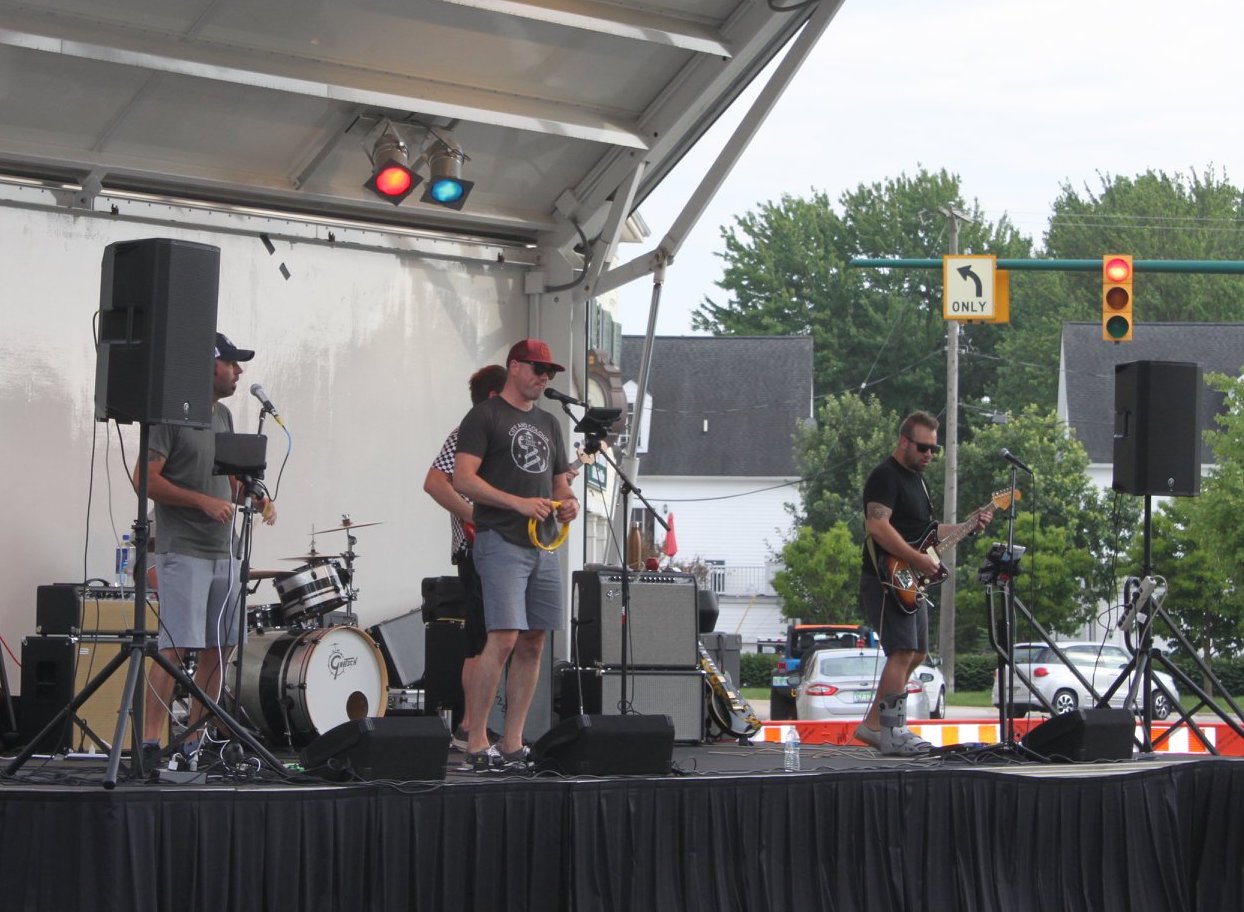 Rally on Ridge Stage Show - Social House News Band performing on an outdoor stage under a canopy: drummer in back, two guitarists and a vocalist with a red cap, with amplifiers and microphones in use.