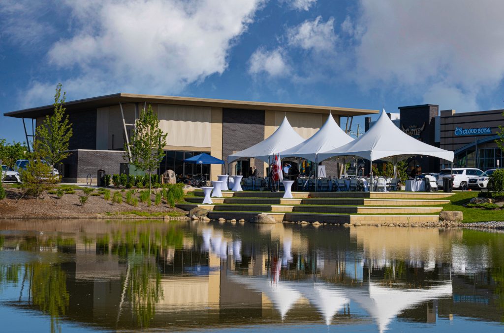 Outdoor event setup with white tents in front of a modern brick building beside a calm pond.
