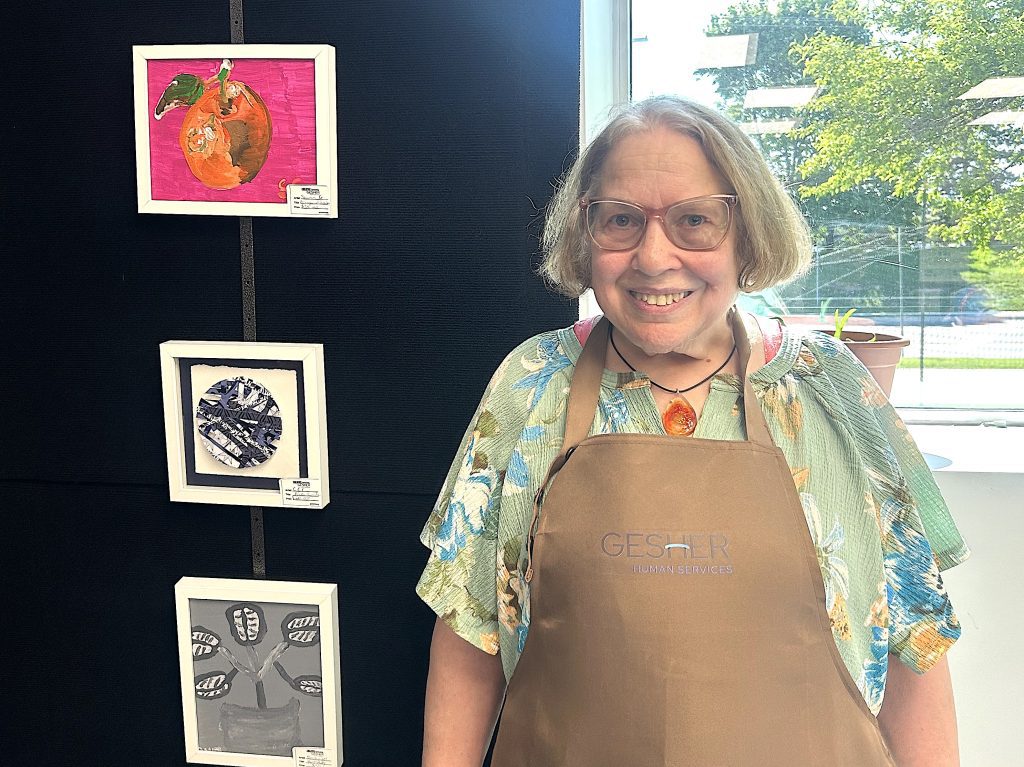 Smiling woman in a floral shirt and brown apron stands beside framed artwork mounted on a black wall in an art display area, window to the right.