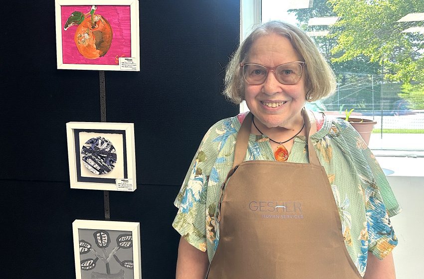 Smiling woman in a floral shirt and brown apron stands beside framed artwork mounted on a black wall in an art display area, window to the right.