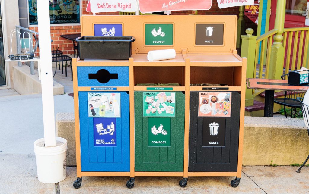 Outdoor three-bin waste sorting station with blue (mixed recyclables), green (compost), and black (waste) bins on wheels; a roll of paper sits on top.