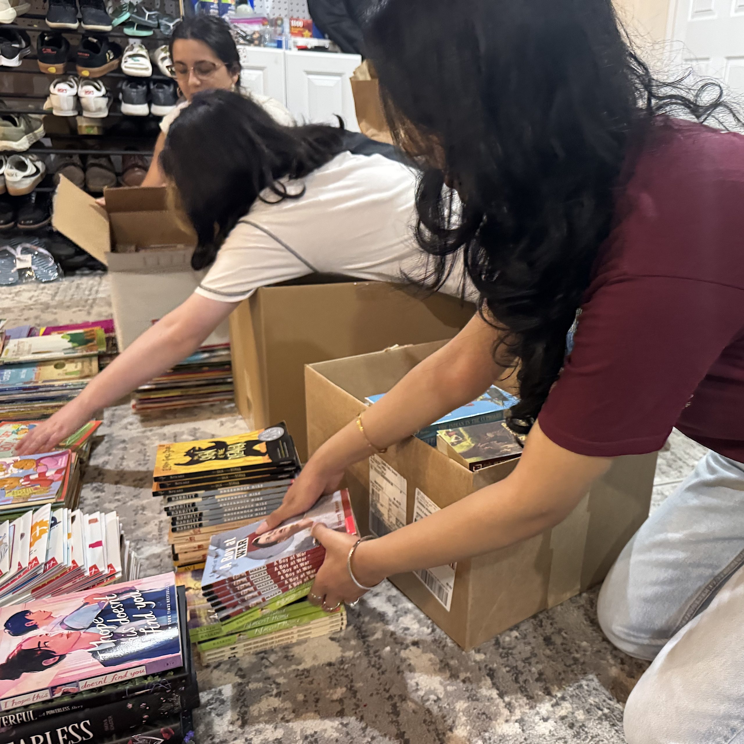 Two women bend over stacks of colorful books on the floor in a bookstore, with a shoe display and cardboard boxes in the background.