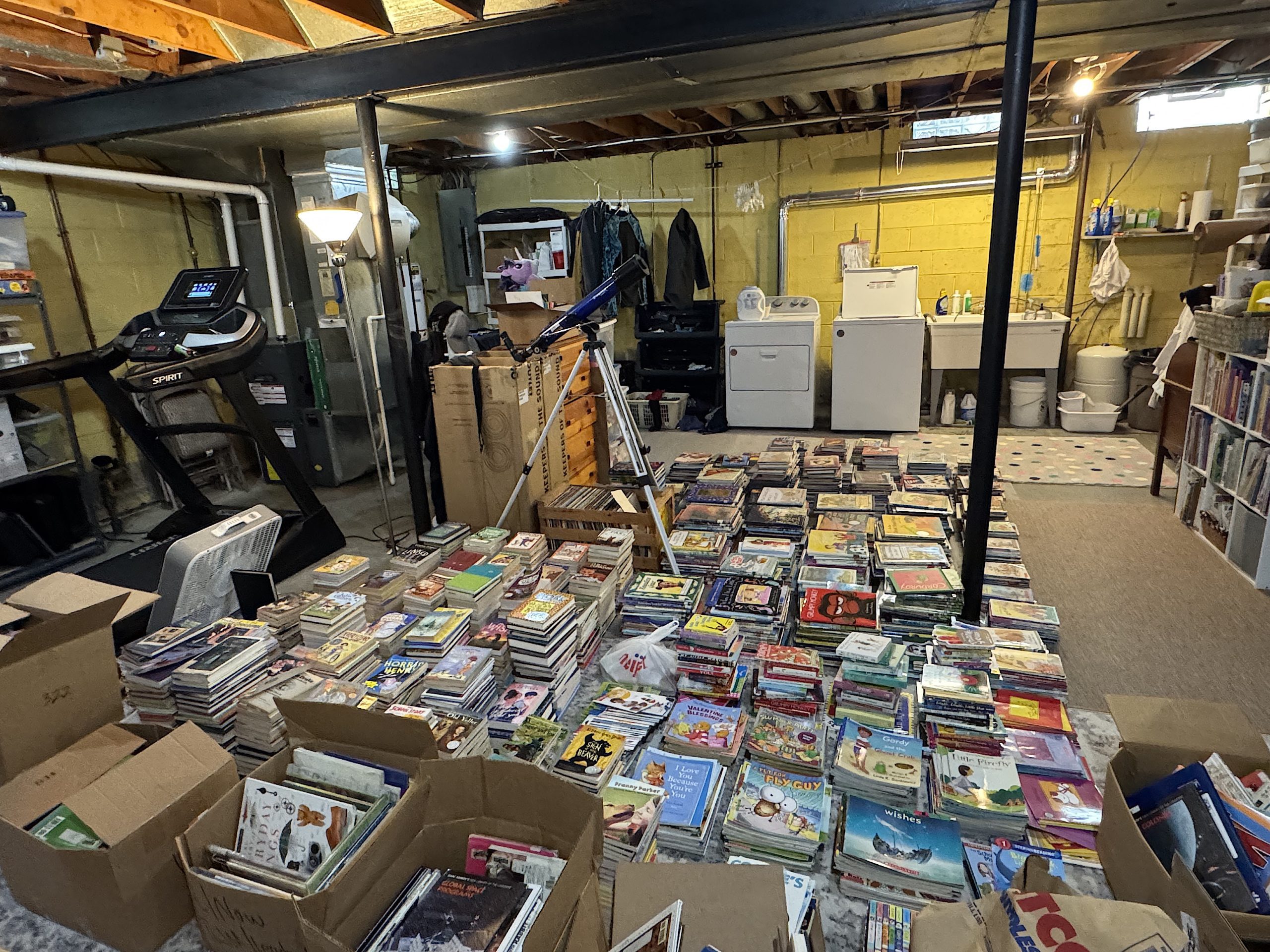 Basement storage area with piles of books and magazines spread across the floor, boxes nearby, and a laundry area in the back corner.