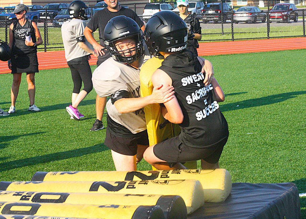 Two players in helmets tackle near padded barriers on a green turf field during a flag football game.