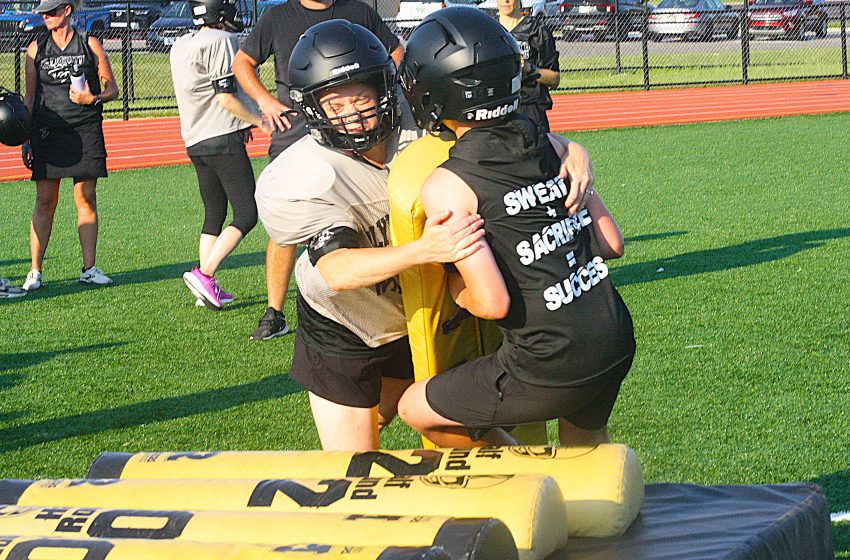 Two players in helmets tackle near padded barriers on a green turf field during a flag football game.