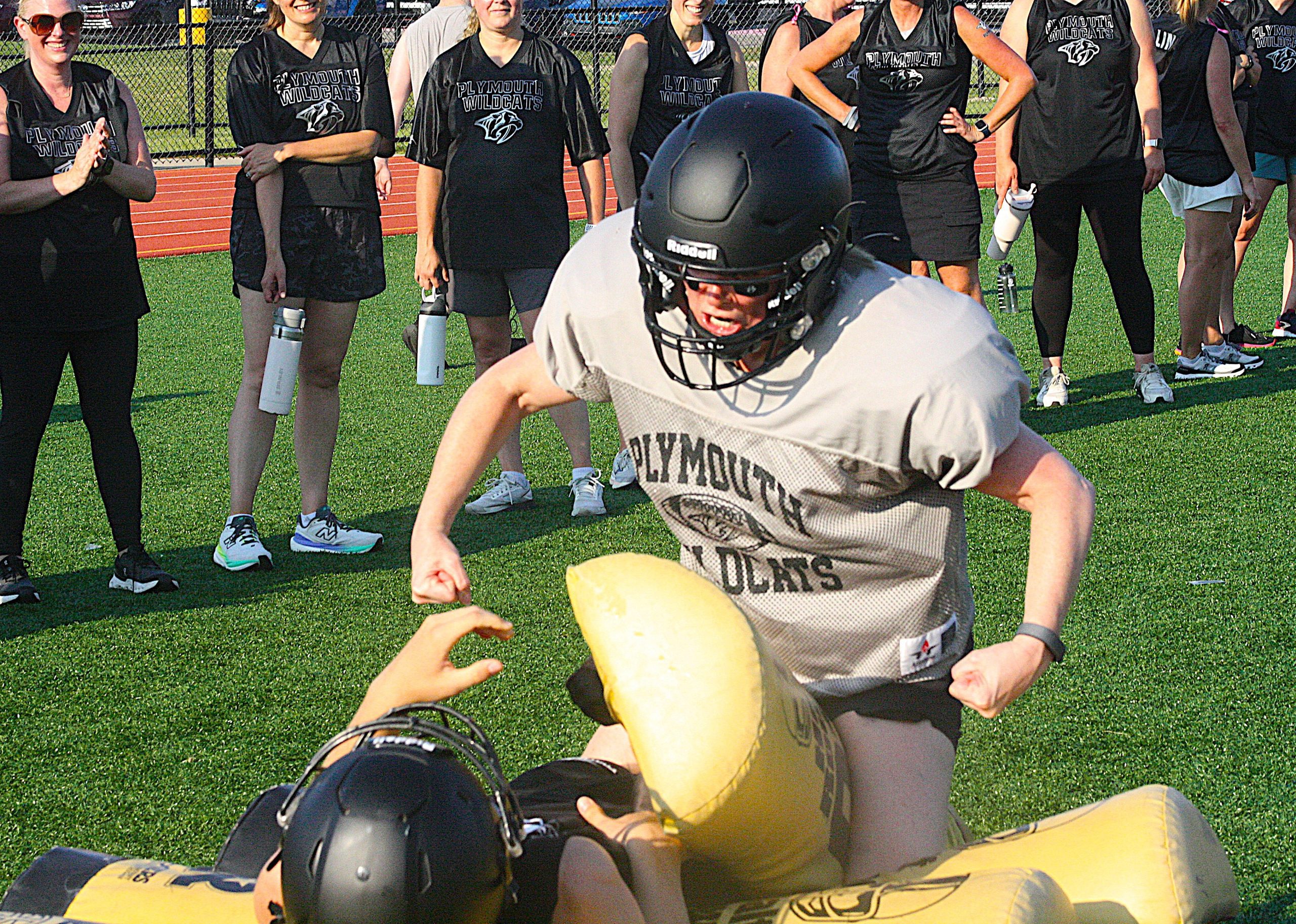 momstackle2 - Social House News Group of Plymouth Wildcats players in black shirts watch a football drill on a grassy field while a teammate in a helmet and white jersey advances toward a yellow tackling pad/o-ling dummy at the foreground
