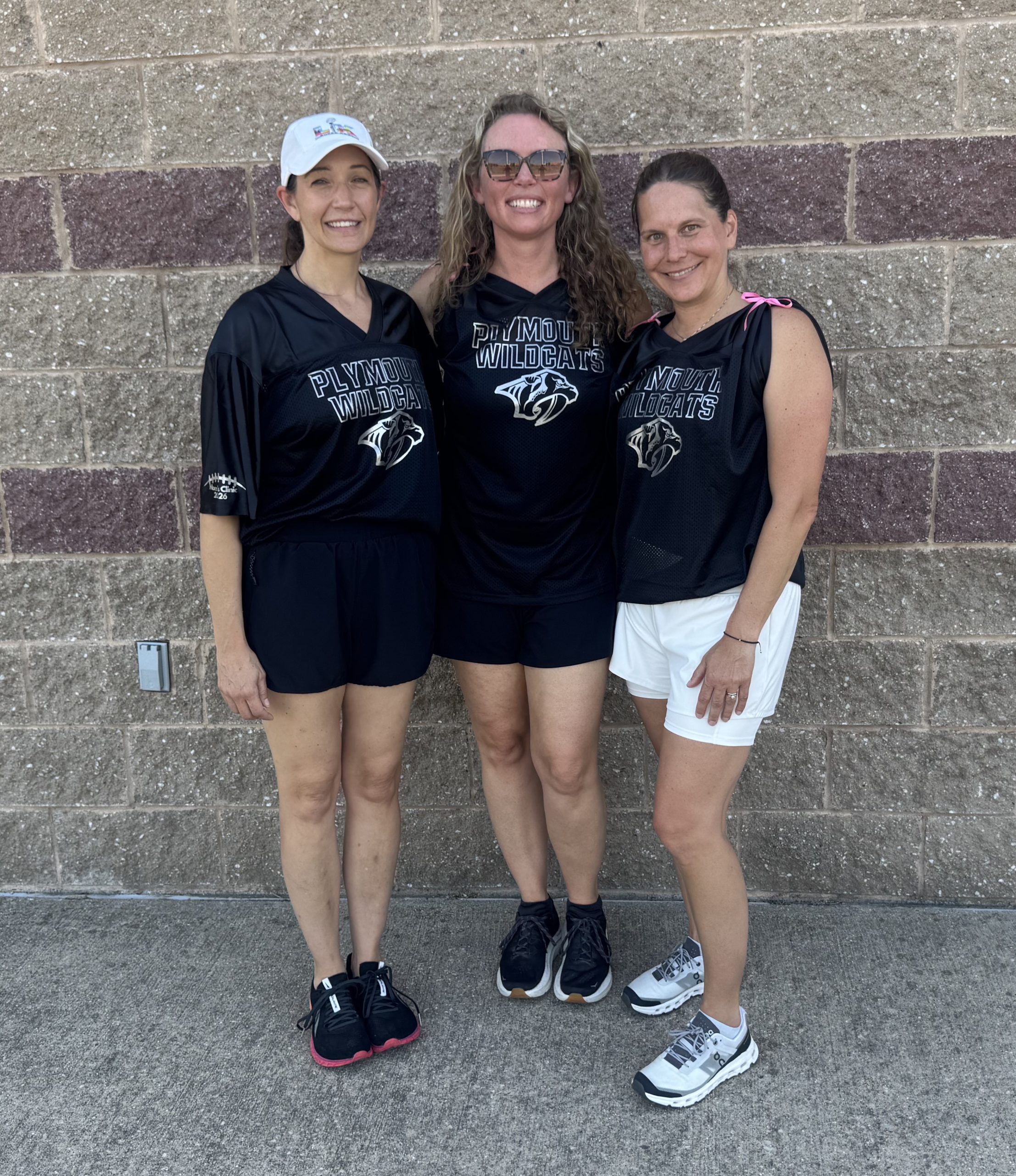 momstackle8 - Social House News Three women in black Plymouth Wildcats jerseys pose together against a brick wall, smiling at the camera.