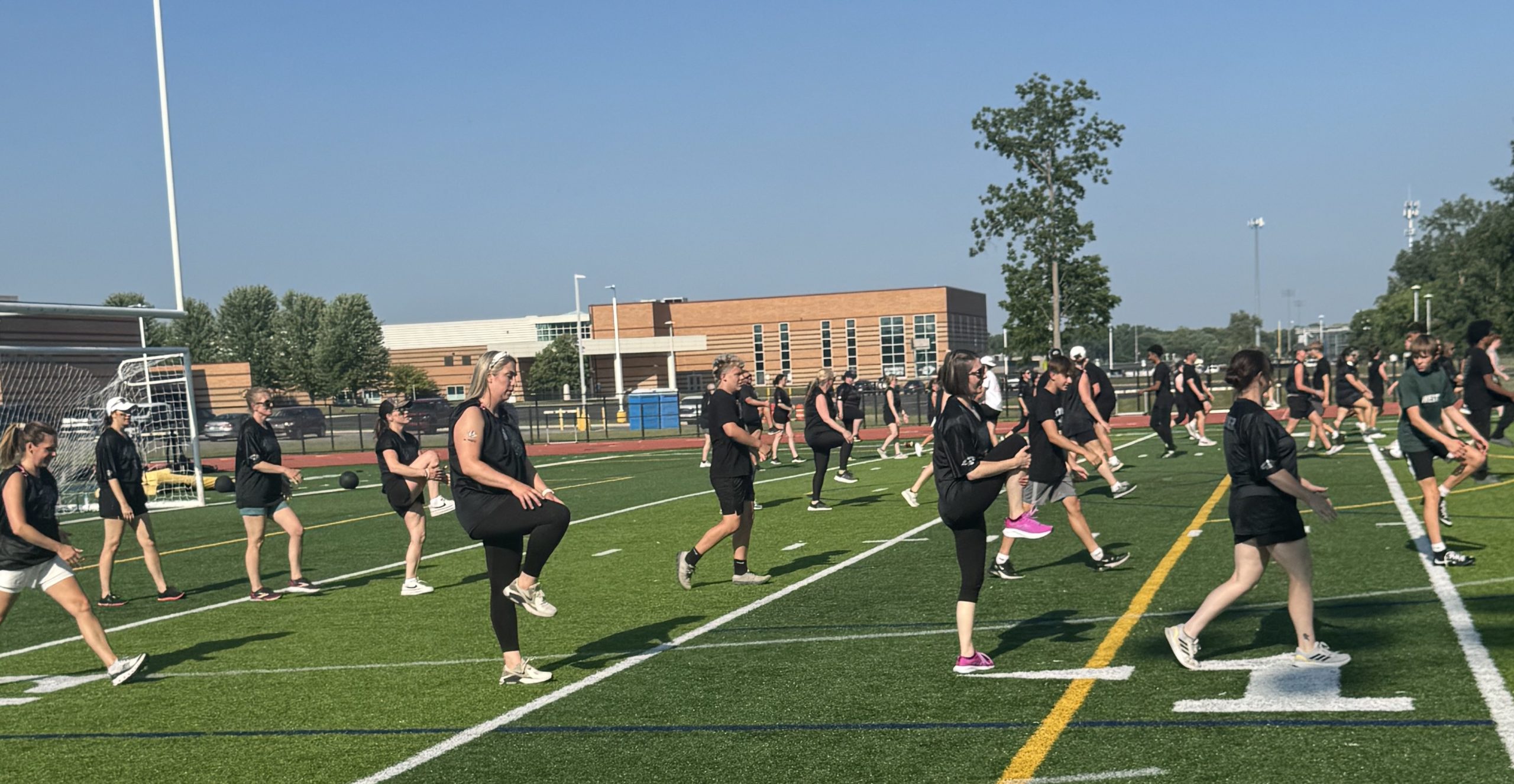 momstackle9 - Social House News Group of people in black athletic outfits warming up on a football field with a goal and school buildings in the background