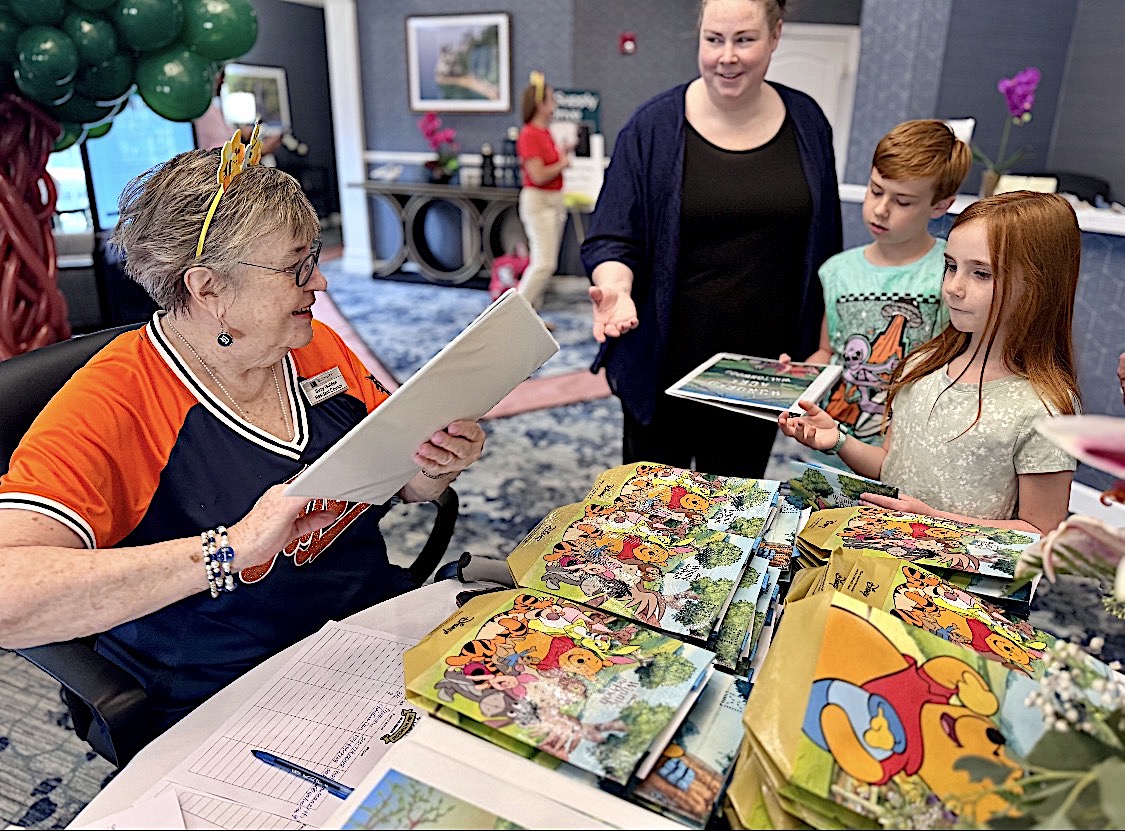 Older woman with glasses in an orange shirt seated at a desk, signing a book for two children and an adult nearby, with colorful children's books on the table.