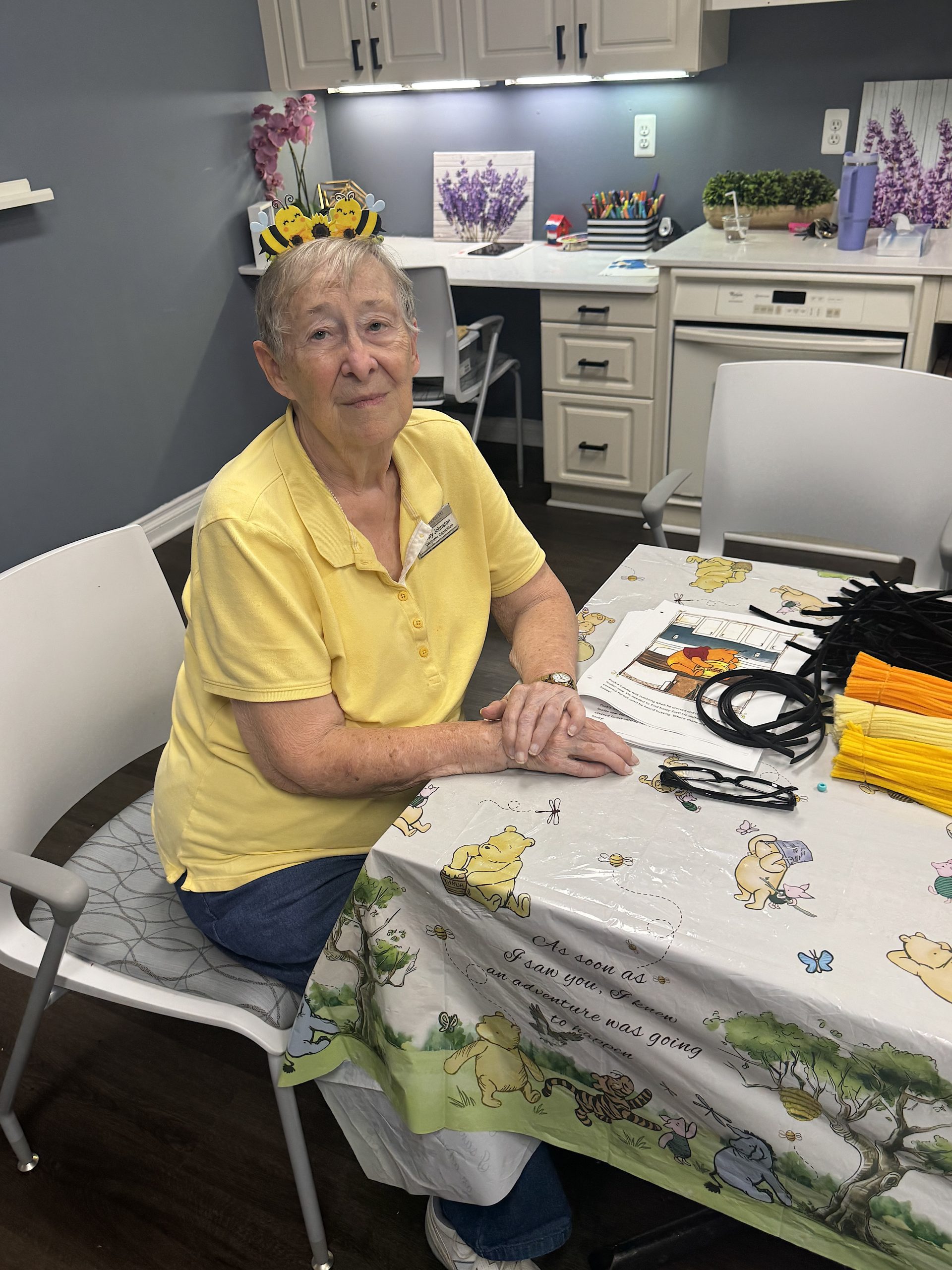 Older woman wearing a yellow polo and a bee headband sits at a craft table in a lavender-themed room with cabinets and lavender decor behind her.