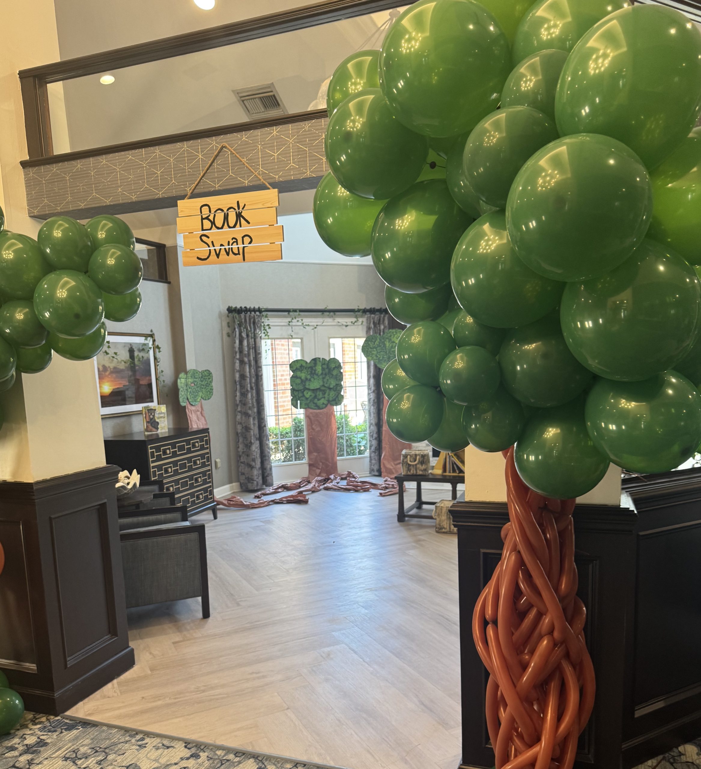 Indoor lobby or shop area with large green balloon columns tied to a dark wood pillar and a hanging wooden sign reading 'Book Swap' in the background.