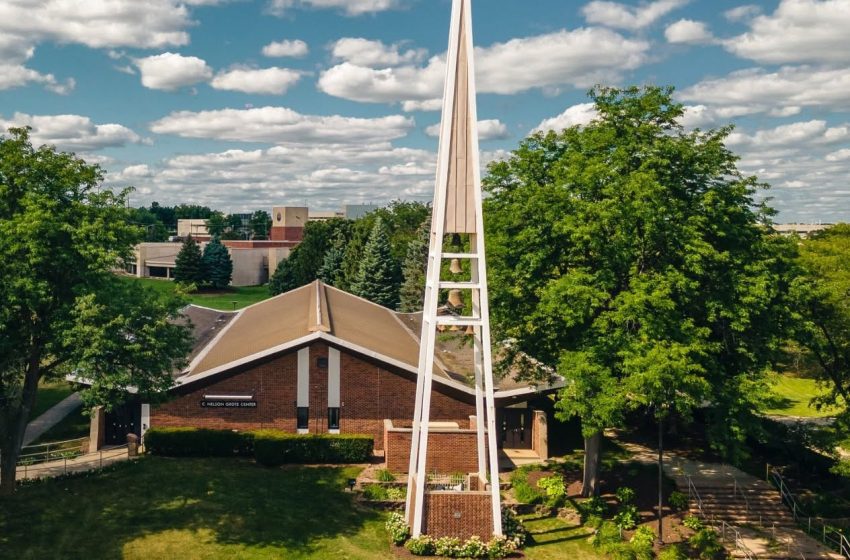 Brick church on a campus with a tall white steeple and bell tower, surrounded by green trees and a blue sky.