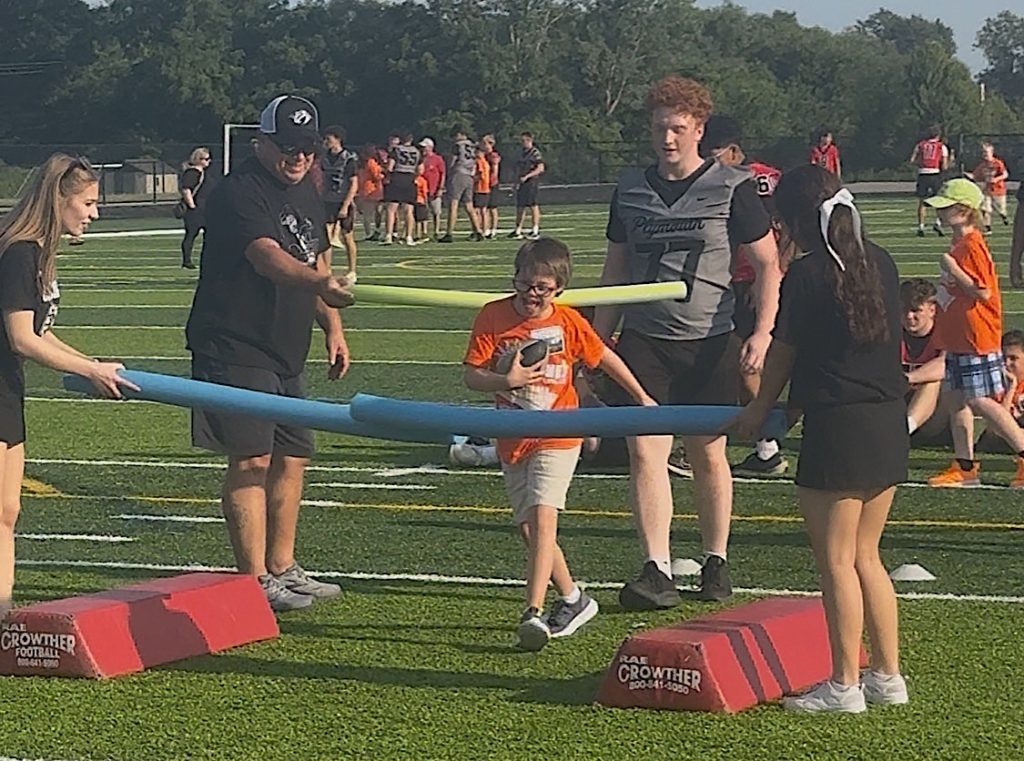 Boy in orange shirt walks under a blue foam baton held by adults and kids on a grassy football field during an outdoor activity.