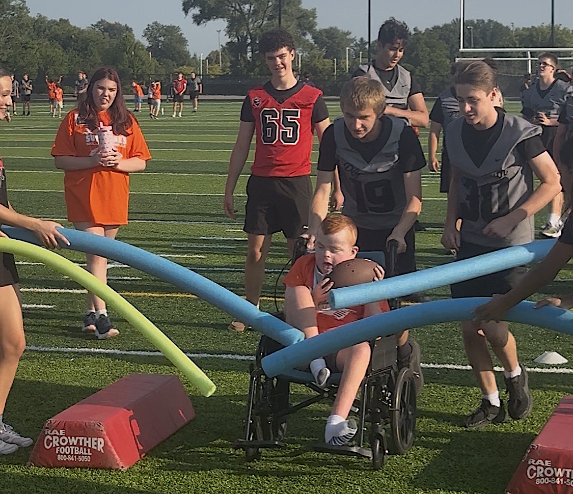 A young boy in a wheelchair moves through a foam-pool-noodle obstacle course on a football field, teammates guiding him nearby.