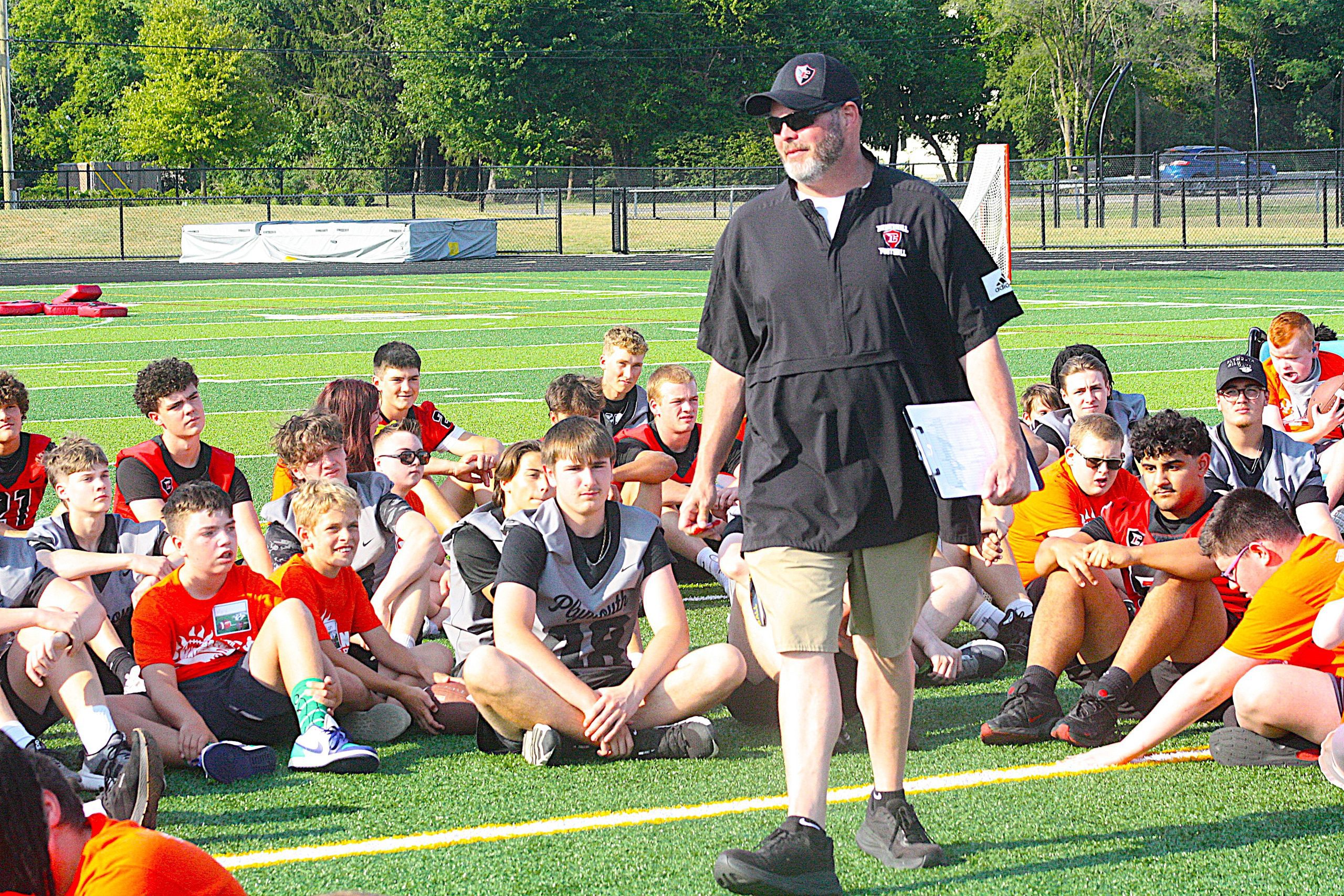 Coach with clipboard addressing a group of youth football players on a sunny field.
